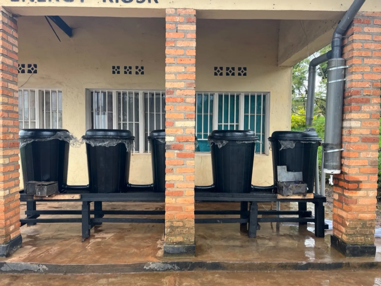 Five black Amazi tanks connected in a row on a raised stand beside a brick building, fed by a downpipe